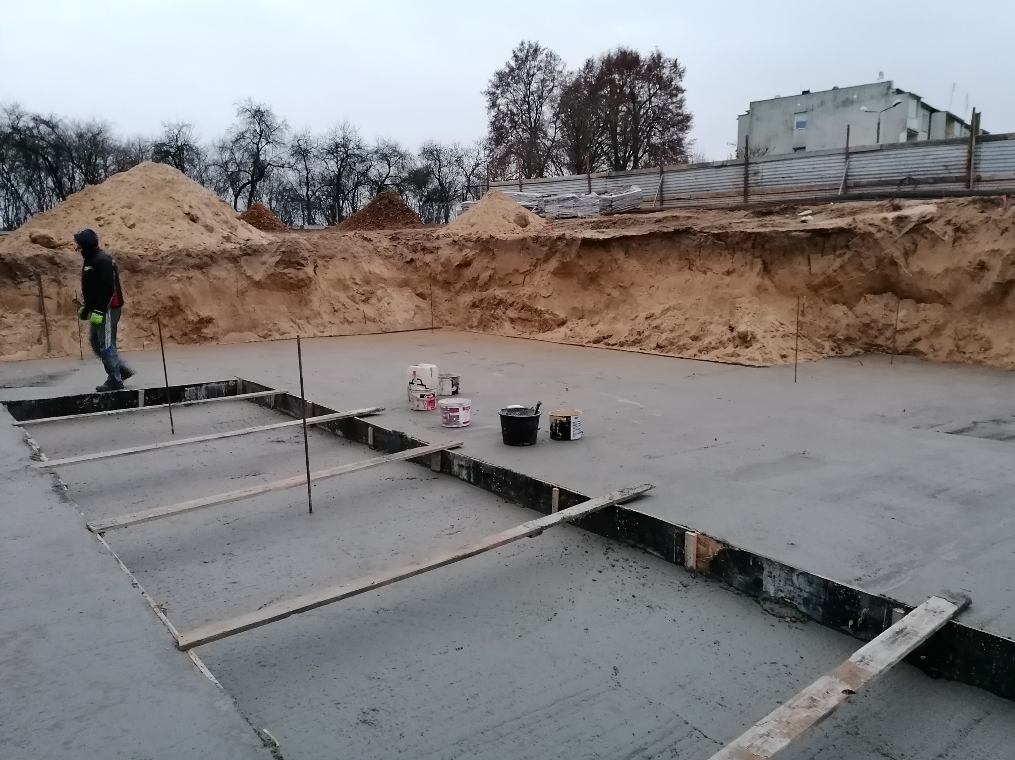 Wylany fundament z deskowaniem i wystającymi prętami zbrojeniowymi, obok leżą puszki z farbą, w tle wykop ziemny i sterta piachu, po lewej stronie przechodzi osoba w odzieży roboczej.
