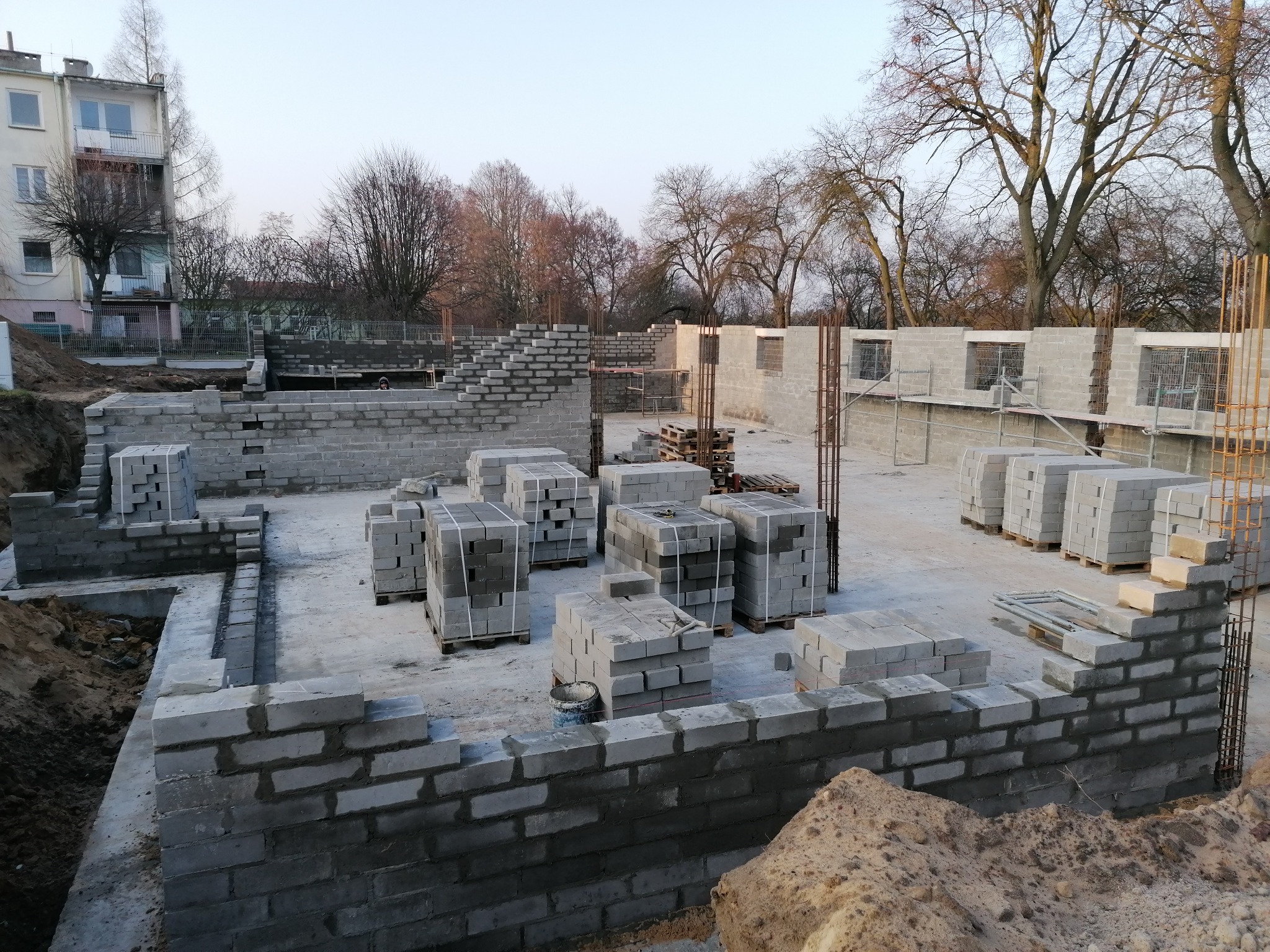 Budowa domu jednorodzinnego w Płocku na etapie fundamentów i wznoszenia ścian z bloczków betonowych. Widoczne zbrojenia, palety z materiałami i wykop pod budynek.
