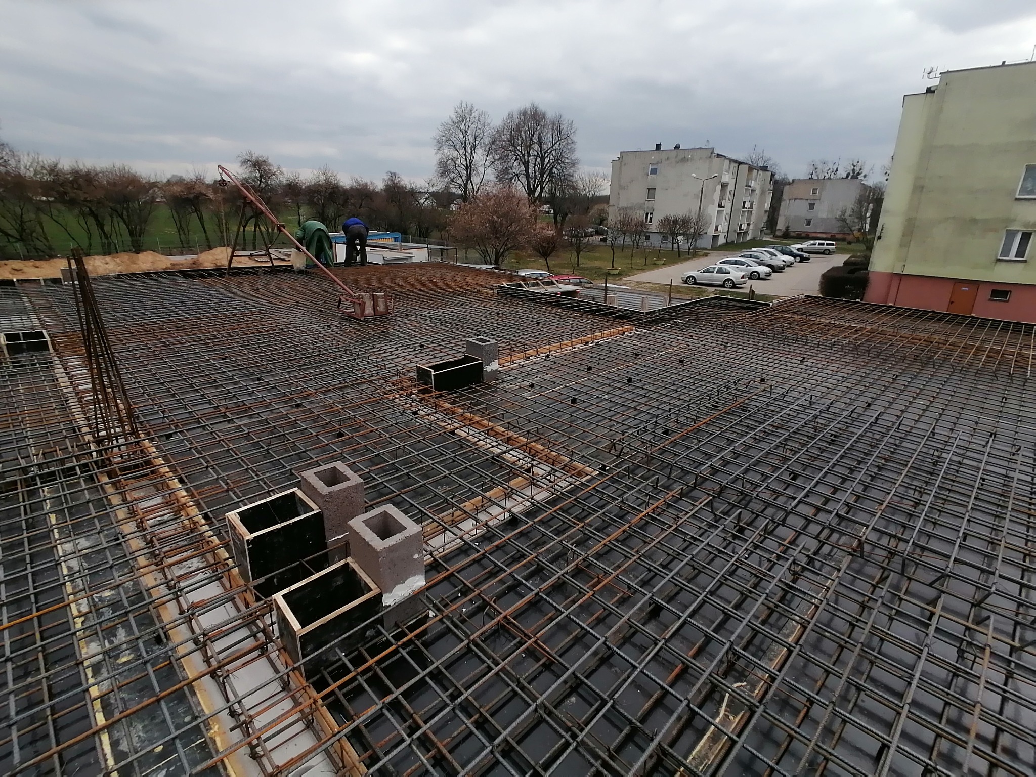 Zbrojenie stropu na budowie, widoczne szalunki i pracownicy w oddali, w tle budynki mieszkalne i pochmurne niebo.