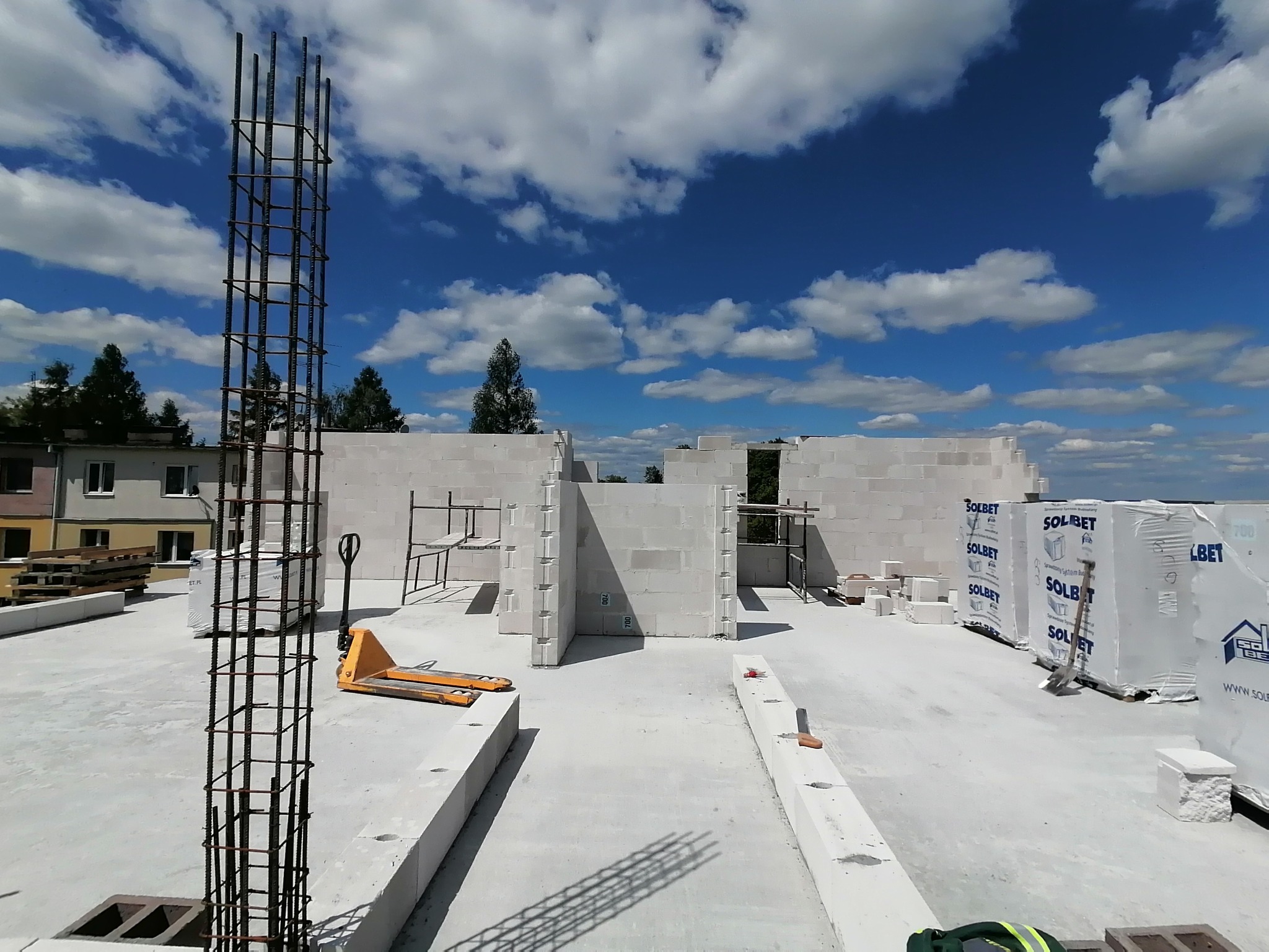 Wznoszenie ścian z bloczków Solbet na budowie, widoczny zbrojony filar, paleciak i niebieskie niebo z chmurami w tle.