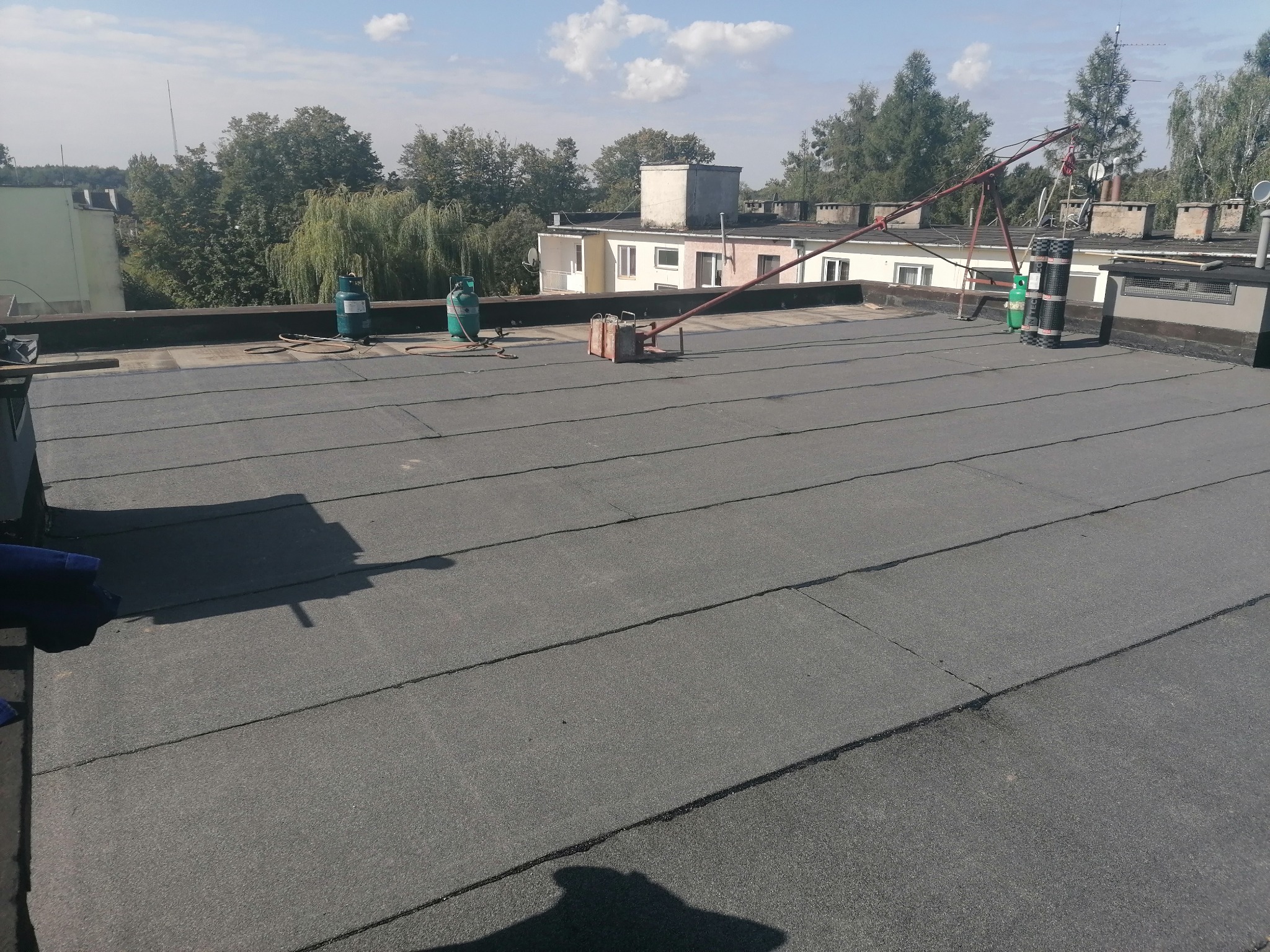 Wykończony, płaski dach pokryty papą termozgrzewalną z widocznymi butlami gazowymi, narzędziami i dźwigiem do transportu materiałów, w tle zabudowa miejska i drzewa.