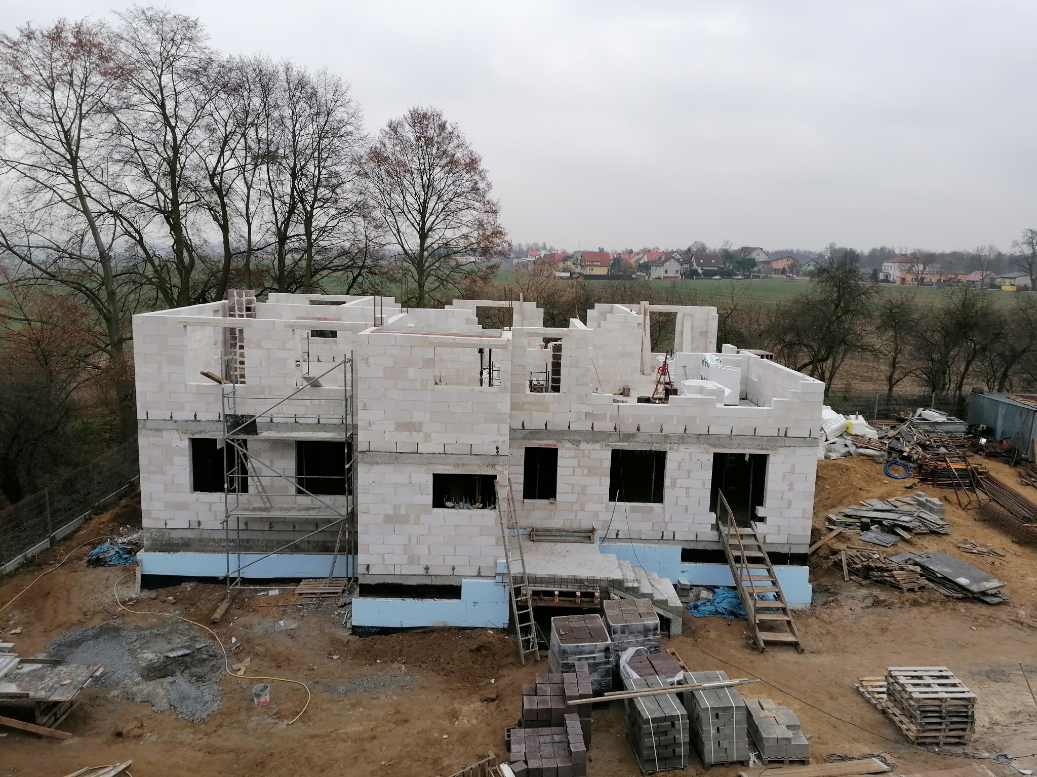 Wznoszenie dwukondygnacyjnego domu z bloczków białego betonu komórkowego, widoczne rusztowania, podłoże ocieplone niebieskim styropianem, w tle zabudowa wiejska i pochmurne niebo.
