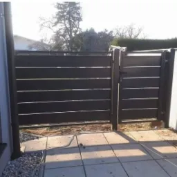 Nowoczesna, pozioma, panelowa brama ogrodzeniowa w kolorze antracytowym, z widocznym zamkiem, zamontowana na betonowej kostce brukowej, z elementami instalacji elektrycznej na ziemi.