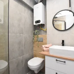 Nowoczesna łazienka z kabiną prysznicową, podwieszaną toaletą, umywalką nablatową na białej szafce z drewnianym blatem i okrągłym lustrem, oświetlona czarnymi lampami punktowymi