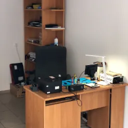 Stanowisko pracy serwisanta komputerowego w Słubicach. Na biurku narzędzia, lampka, monitor, głośnik, mata serwisowa, obudowa komputera obok biurka, półka z akcesoriami w tle.