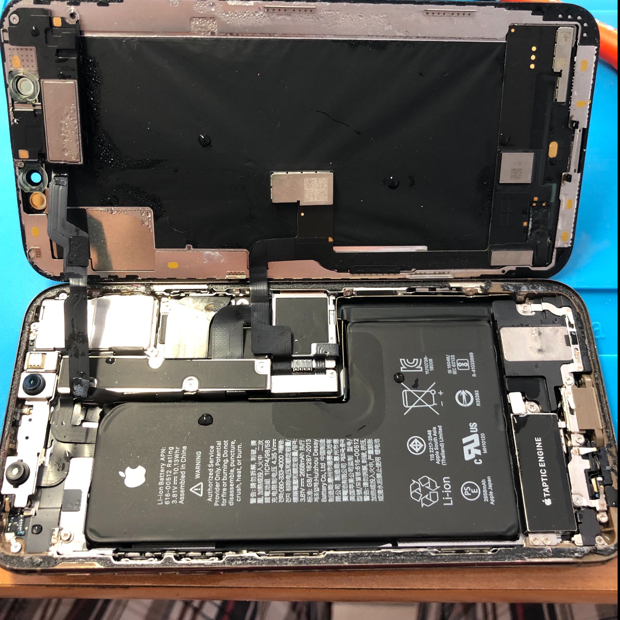 Rozłożony iPhone X z widoczną baterią i elementami wewnętrznymi, krople cieczy na ekranie, ukazujący proces naprawy lub demontażu.