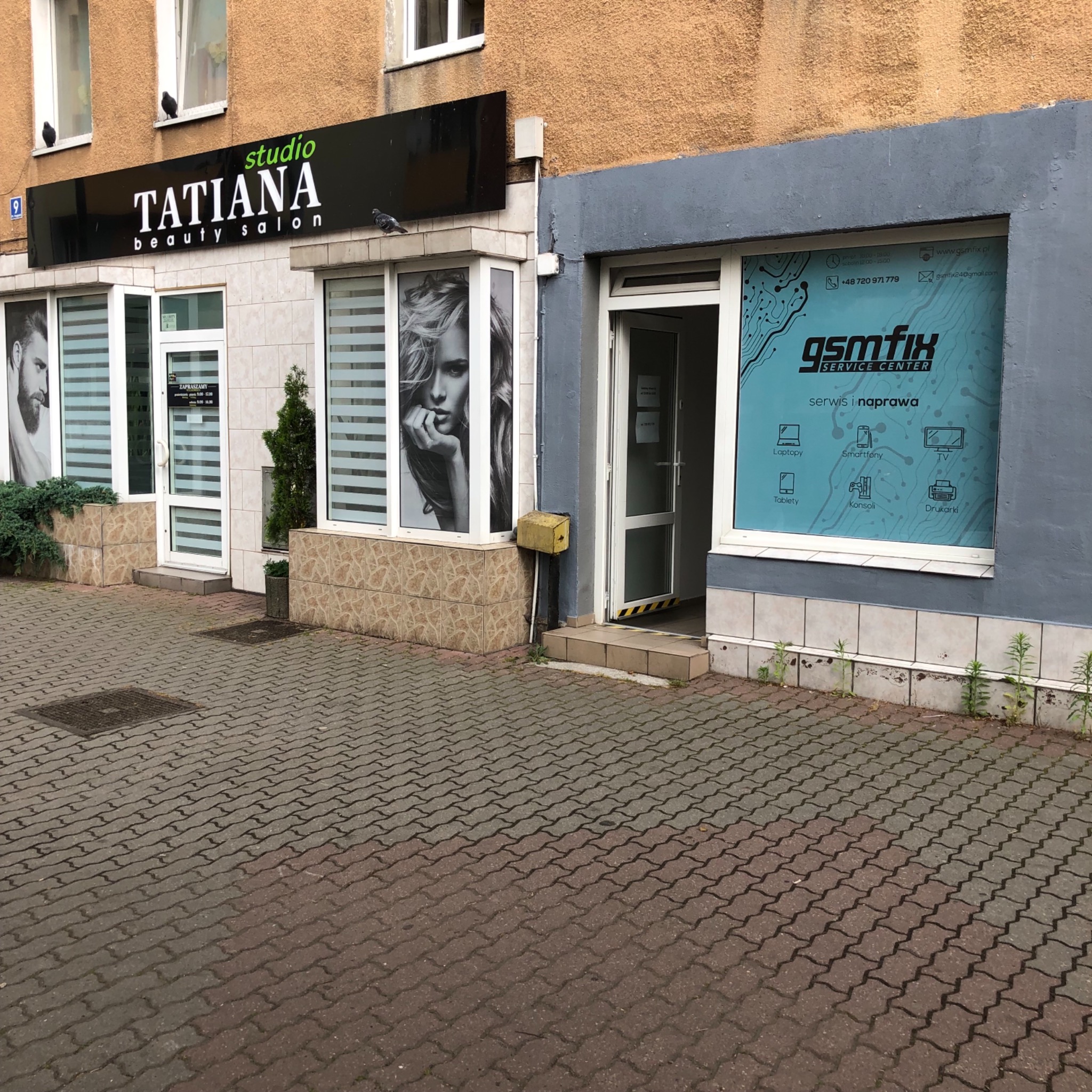 Fasada budynku z dwoma lokalami: salon urody Tatiana z lewej i serwis GSMfix z prawej, z reklamami w oknach i wejściami z poziomu chodnika wyłożonego kostką.