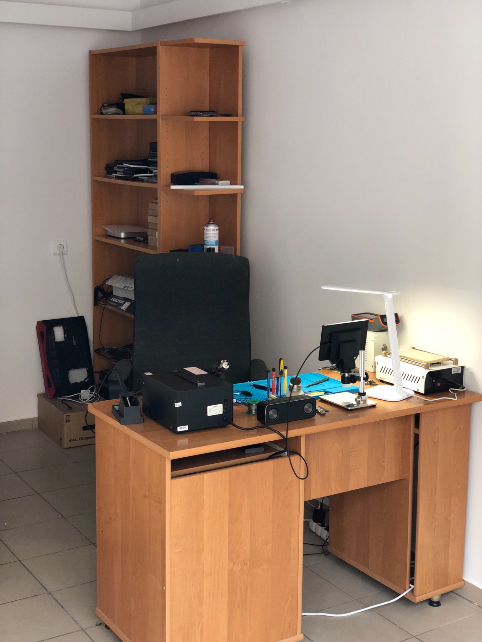 Stanowisko pracy serwisanta komputerowego w Słubicach. Na biurku narzędzia, lampka, monitor, głośnik, mata serwisowa, obudowa komputera obok biurka, półka z akcesoriami w tle.