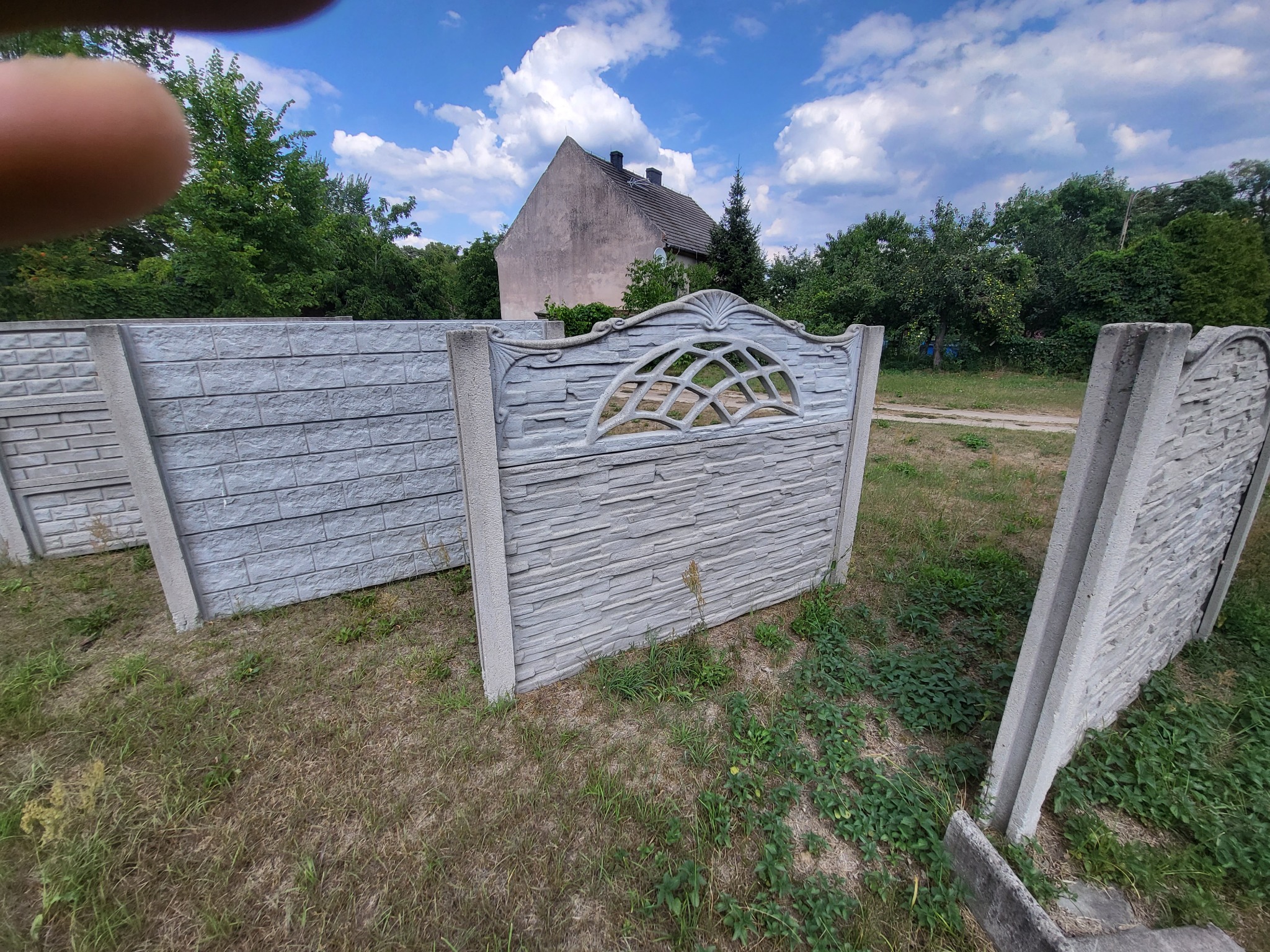 Fragmenty szarego, betonowego ogrodzenia z dekoracyjnym panelem, ustawione na trawie przed domem w słoneczny dzień.