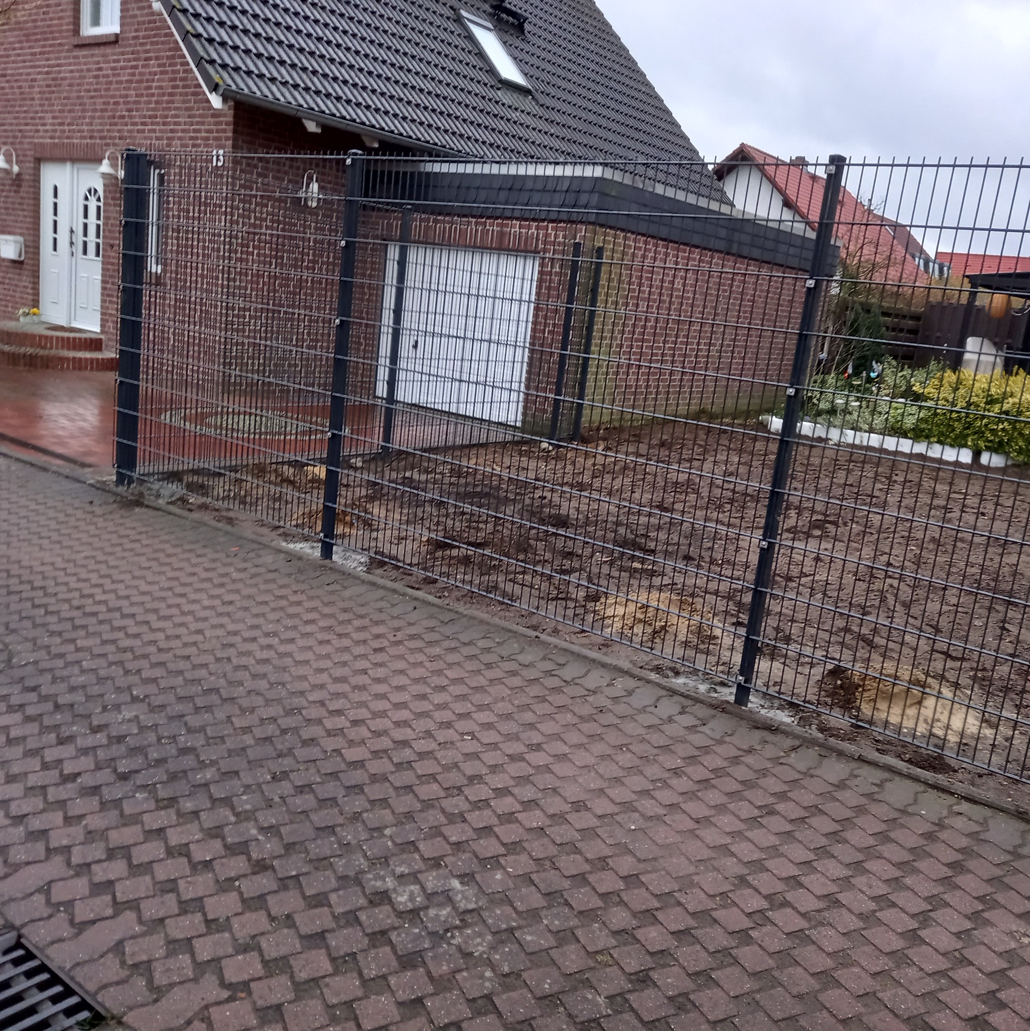 Montaż ogrodzenia panelowego z drutu na posesji z domem z czerwonej cegły i garażem, widok z chodnika z kostki.