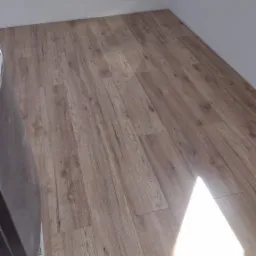 Wykończona podłoga z paneli imitujących drewno, świeżo po położeniu, z widocznym odblaskiem światła oraz fragmentem opakowania po materiałach budowlanych w rogu.