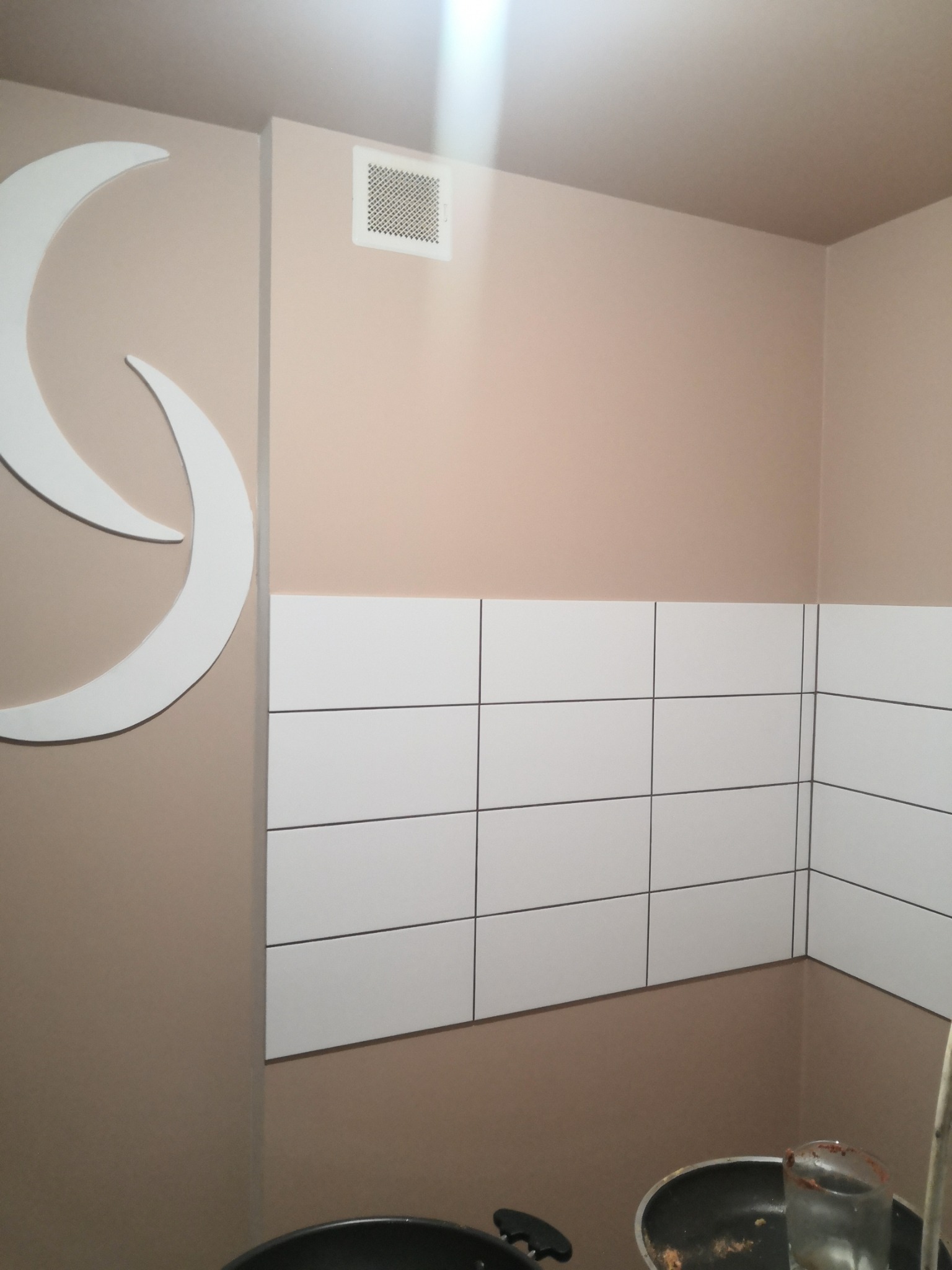 Beżowa ściana z dekoracją w kształcie księżyca, wentylacją i białymi płytkami w kuchni, widoczne naczynia.