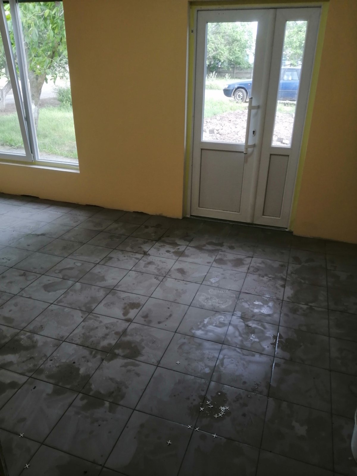 Świeżo ułożone, ciemnoszare płytki podłogowe w pomieszczeniu z żółtymi ścianami, widoczne krzyżyki dystansowe i ślady po fugowaniu, okno i drzwi balkonowe z widokiem na zewnątrz.