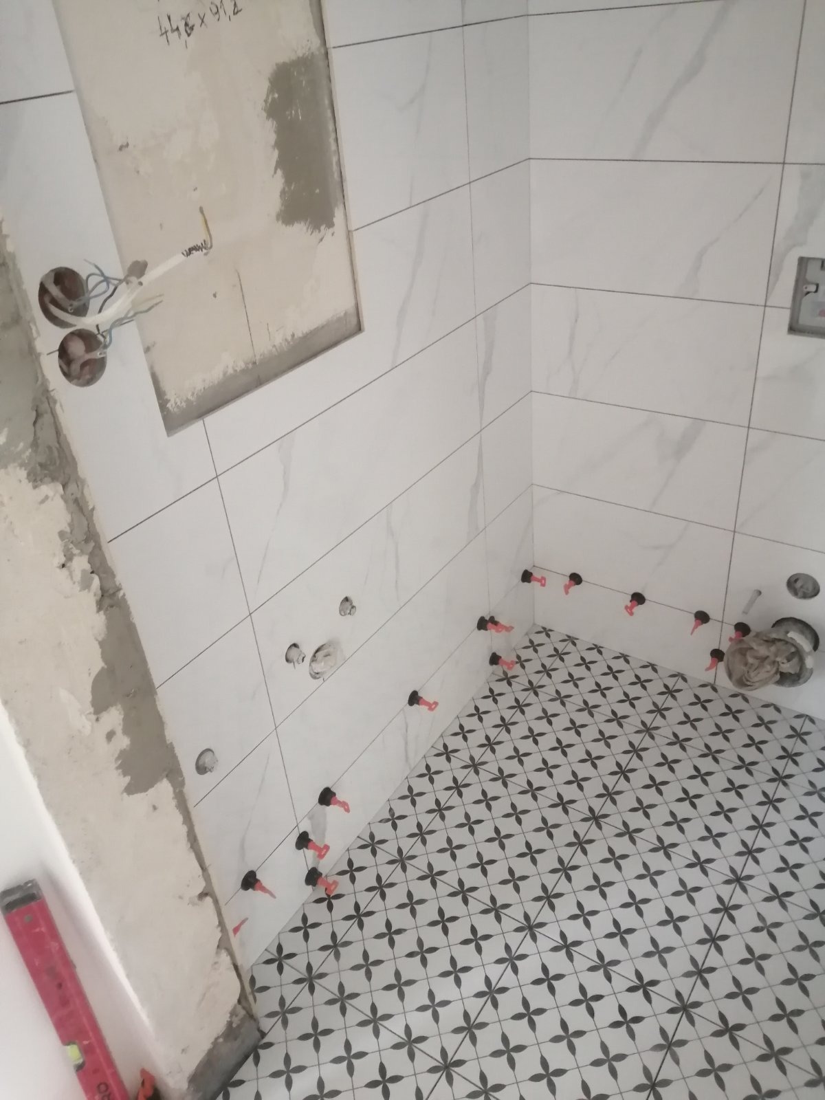 Układanie białych płytek imitujących marmur na ścianach i czarno-białych płytek w kwiatowy wzór na podłodze w łazience, z użyciem poziomicy i systemu poziomowania płytek z czerwonymi klinami.