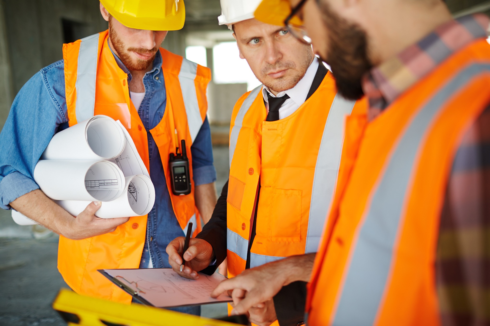 Trzech mężczyzn w kamizelkach odblaskowych i kaskach omawia plany na budowie w Bielsku-Białej. Jeden z nich, w garniturze, nanosi poprawki na rysunek.