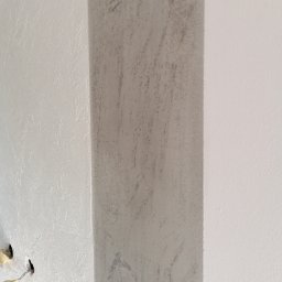 Tomasz www - Fragment ściany z dekoracyjną, szarą kolumną o chropowatej fakturze, sąsiadującą z gładką, białą powierzchnią; widoczne otwory po instalacji elektrycznej.