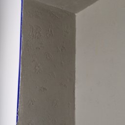Tomasz www - Szara ściana z fakturą, zabezpieczona niebieską taśmą malarską przy białym narożniku, fragment sufitu z tą samą fakturą.