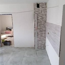 Tomasz www - Wnętrze w trakcie remontu: częściowo wykończona ściana z płytek imitujących cegłę, surowa podłoga z szarych płytek, otwór drzwiowy bez drzwi, widoczne materiały budowlane w tle.