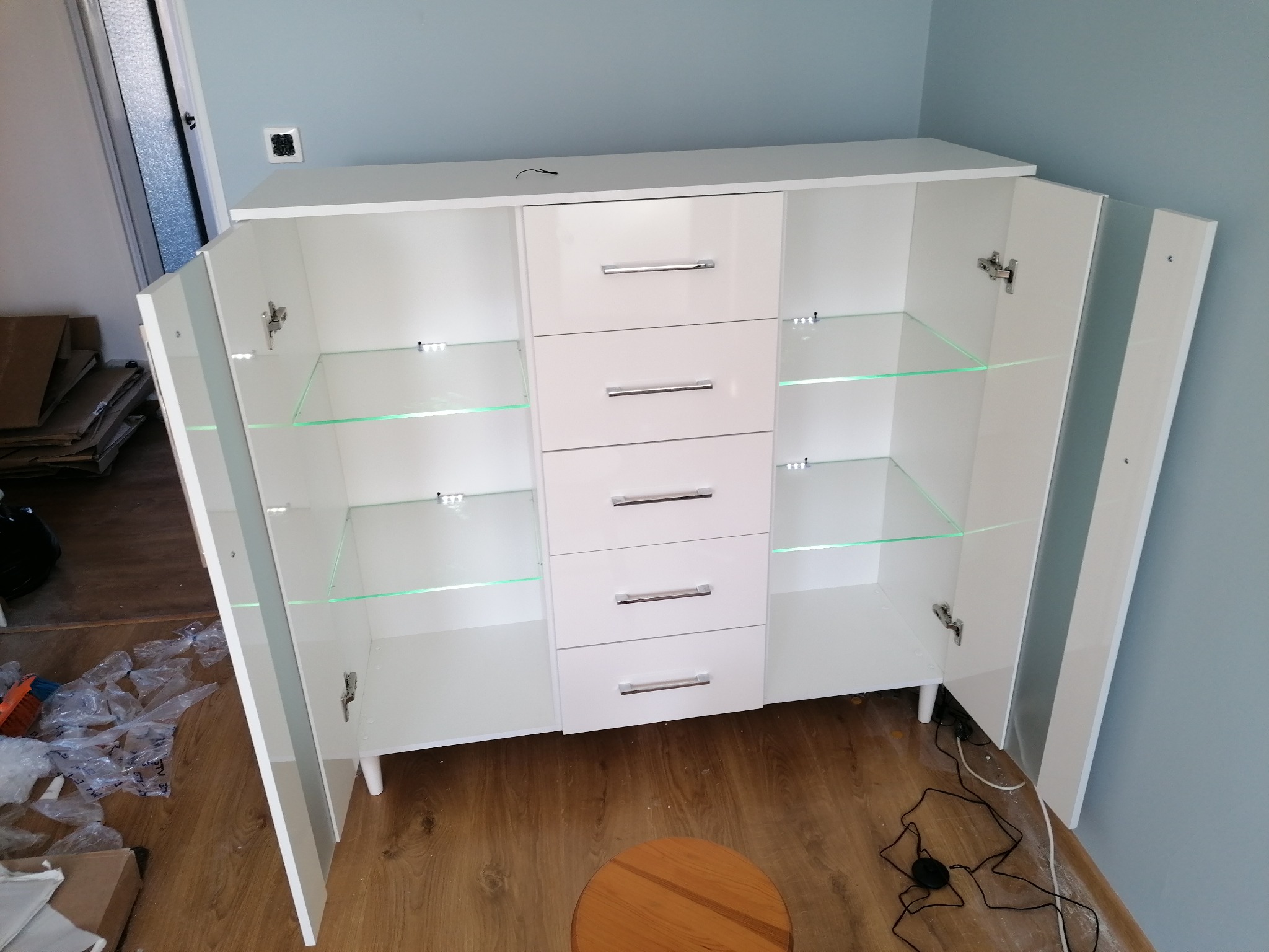 Biała komoda z szufladami i szklanymi półkami, z otwartymi drzwiczkami, widoczna w trakcie montażu oświetlenia LED, Dębica.