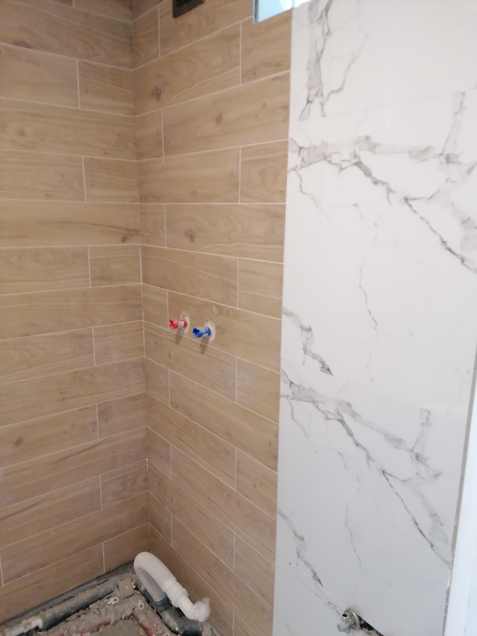 Fragment łazienki w trakcie wykańczania, z położonymi płytkami imitującymi drewno na jednej ścianie i białymi płytkami z marmurkowym wzorem na drugiej, widoczne wystające rury z instalacji wodnej.