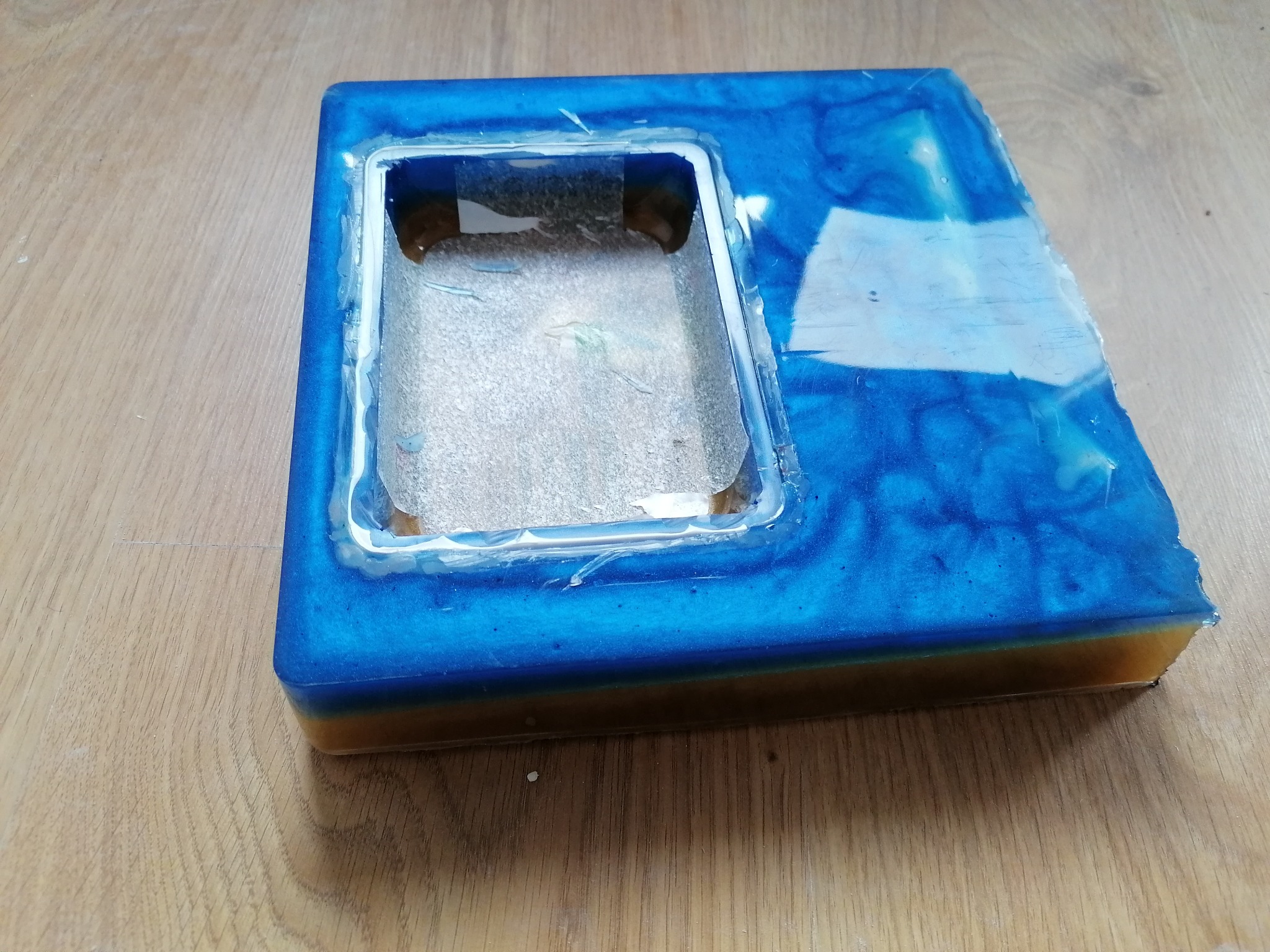 Nieregularny odlew z żywicy epoksydowej w odcieniach błękitu i brązu, z otworem w centrum, leżący na drewnianej powierzchni.