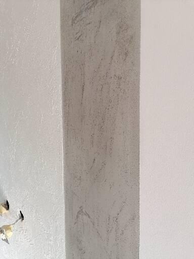 Fragment ściany z widocznym efektem dekoracyjnym w odcieniach szarości, sąsiadujący z gładką, białą powierzchnią. Widoczne gniazdka elektryczne po lewej stronie.