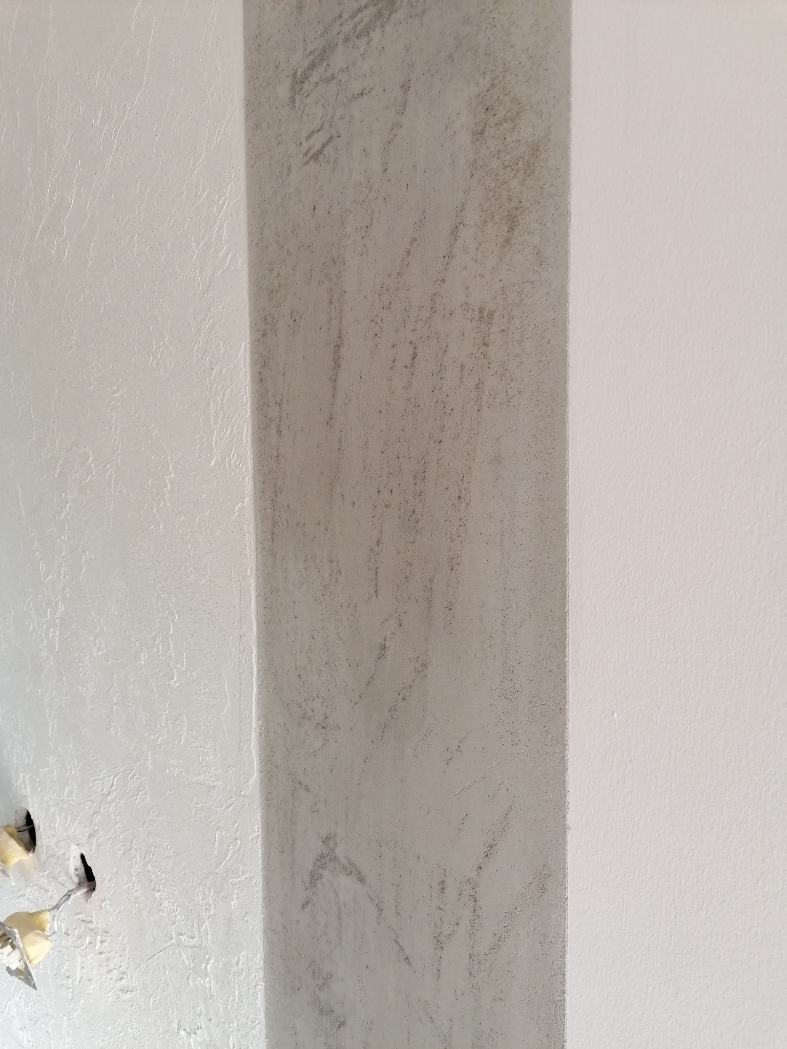 Fragment ściany z dekoracyjną, szarą kolumną o chropowatej fakturze, sąsiadującą z gładką, białą powierzchnią; widoczne otwory po instalacji elektrycznej.