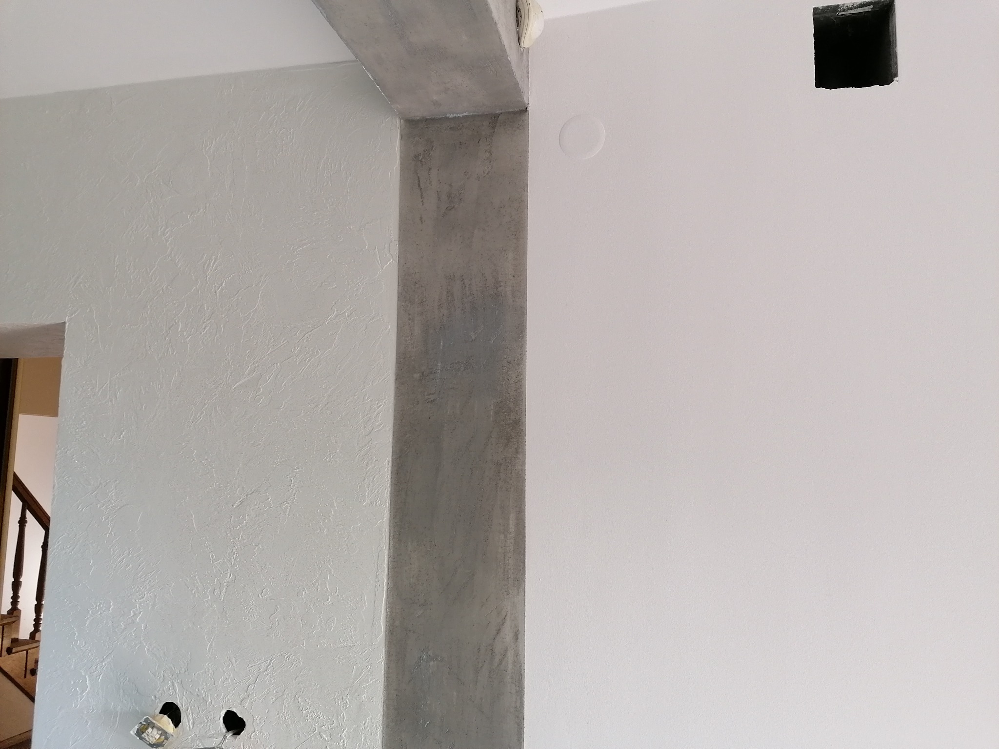 Szara ściana z efektem dekoracyjnym, betonowy filar z widoczną strukturą, otwór w ścianie i wyjścia na kable elektryczne w dolnej części ściany.