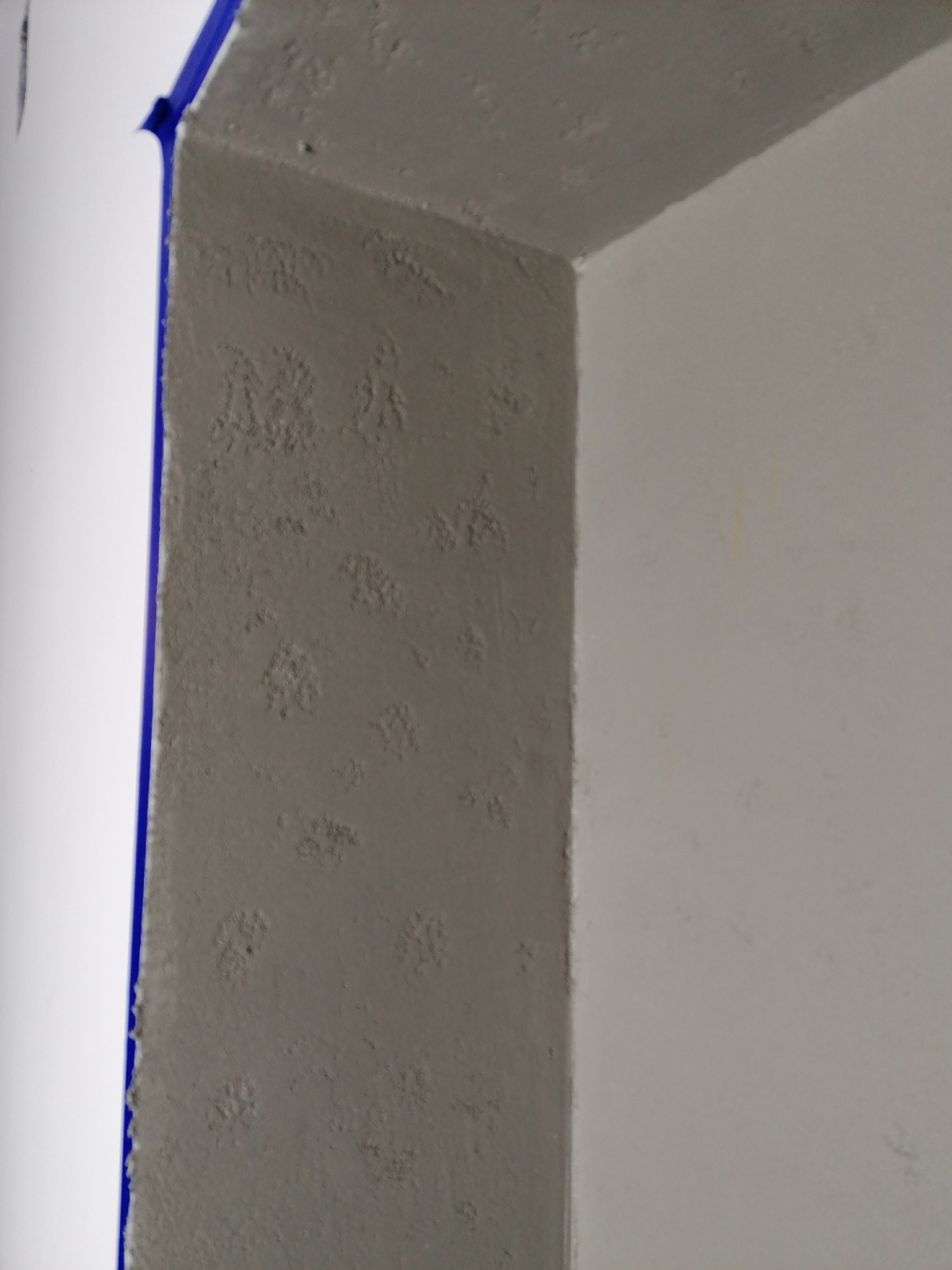 Szara ściana z fakturą, zabezpieczona niebieską taśmą malarską przy białym narożniku, fragment sufitu z tą samą fakturą.