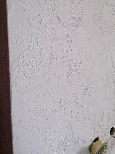 Biała ściana z dekoracyjną fakturą, widoczny fragment brązowej framugi drzwi po lewej stronie i szpachelka w dolnym prawym rogu.