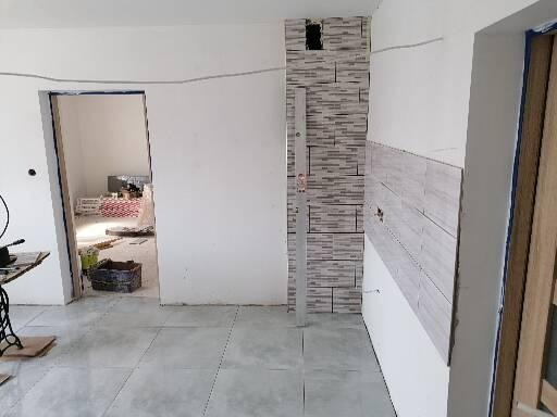 Wnętrze w trakcie remontu: częściowo wykończona ściana z płytek imitujących cegłę, surowa podłoga z szarych płytek, otwór drzwiowy bez drzwi, widoczne materiały budowlane w tle.