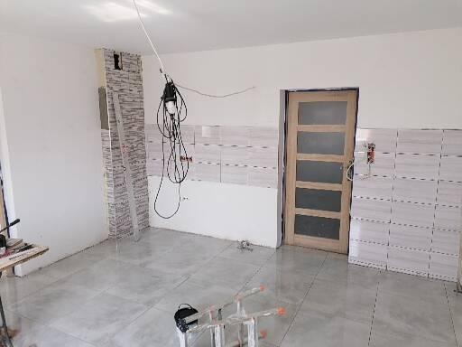 Wnętrze w trakcie remontu: częściowo wykończona ściana z płytek, drewniane drzwi z szybami, drabina, oraz wiszący przewód elektryczny z reflektorem.