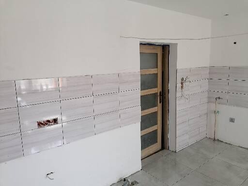 Wnętrze w trakcie remontu: ściany częściowo wyłożone białymi płytkami imitującymi marmur, widoczna drewniana ościeżnica drzwiowa z szybami oraz wystające przewody elektryczne.