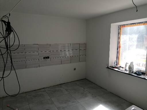 Wnętrze kuchni w trakcie remontu, widoczne ułożone płytki nad blatem, niepodłączone kable elektryczne zwisające ze ściany oraz okno zabezpieczone folią malarską.