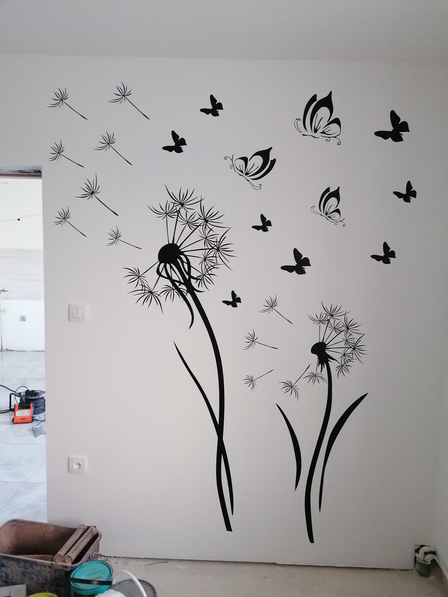 Naklejki ścienne z motywem dmuchawców i motyli na białej ścianie w trakcie remontu, widoczne puszki z farbą i lampa robocza.