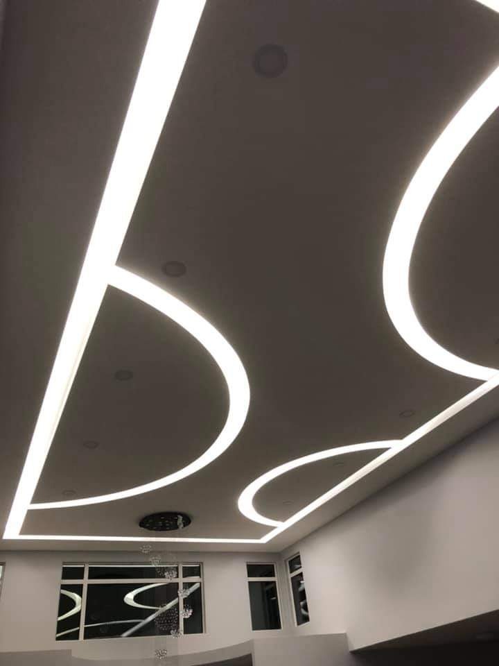 Nowoczesny sufit podwieszany z oświetleniem LED w kształcie łuków, widoczne okna z dekoracyjnymi elementami odbijającymi światło.