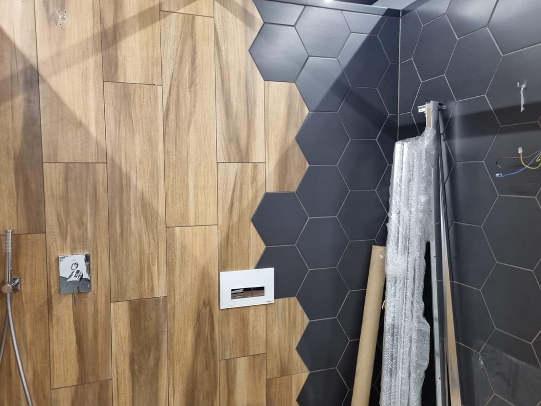Wnętrze łazienki w trakcie wykańczania: połączenie drewnopodobnych płytek ściennych i grafitowych heksagonów, widoczne elementy instalacji prysznicowej i stelaż podtynkowy.