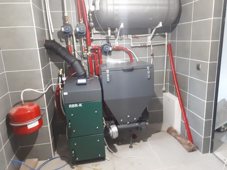 Nowoczesny piec na paliwo stałe RBR-K z zasobnikiem, zainstalowany w kotłowni wyłożonej szarymi płytkami, widoczne rury w kolorze czerwonym i białe elementy instalacji.