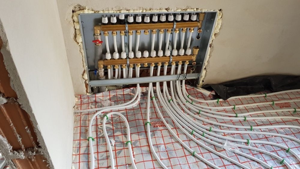 Rozdzielacz ogrzewania podłogowego zamontowany w ścianie, z widocznymi białymi rurkami ułożonymi na folii izolacyjnej z czerwonym nadrukiem, zabezpieczone zielonymi opaskami, surowy stan...
