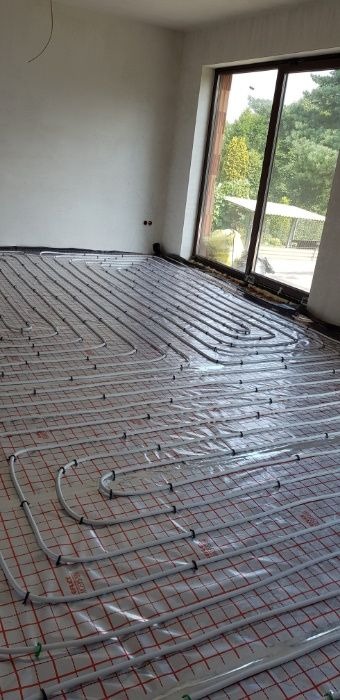 Białe rury ułożone na czerwonej kratownicy podłoża, przygotowane do wylewki ogrzewania podłogowego w jasnym pomieszczeniu z dużym oknem.