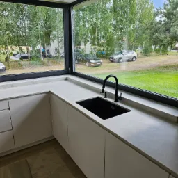 Jasnoszary blat kamienny przy oknie z widokiem na zieleń, czarny zlew i bateria w nowoczesnej kuchni z białymi szafkami.