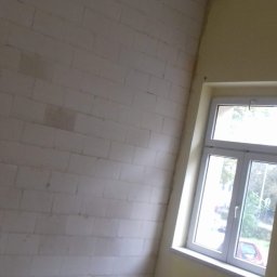 Mirbud - Surowa ściana z białych bloczków w trakcie prac wykończeniowych, widoczne okno i sufit z lampą rastrową.