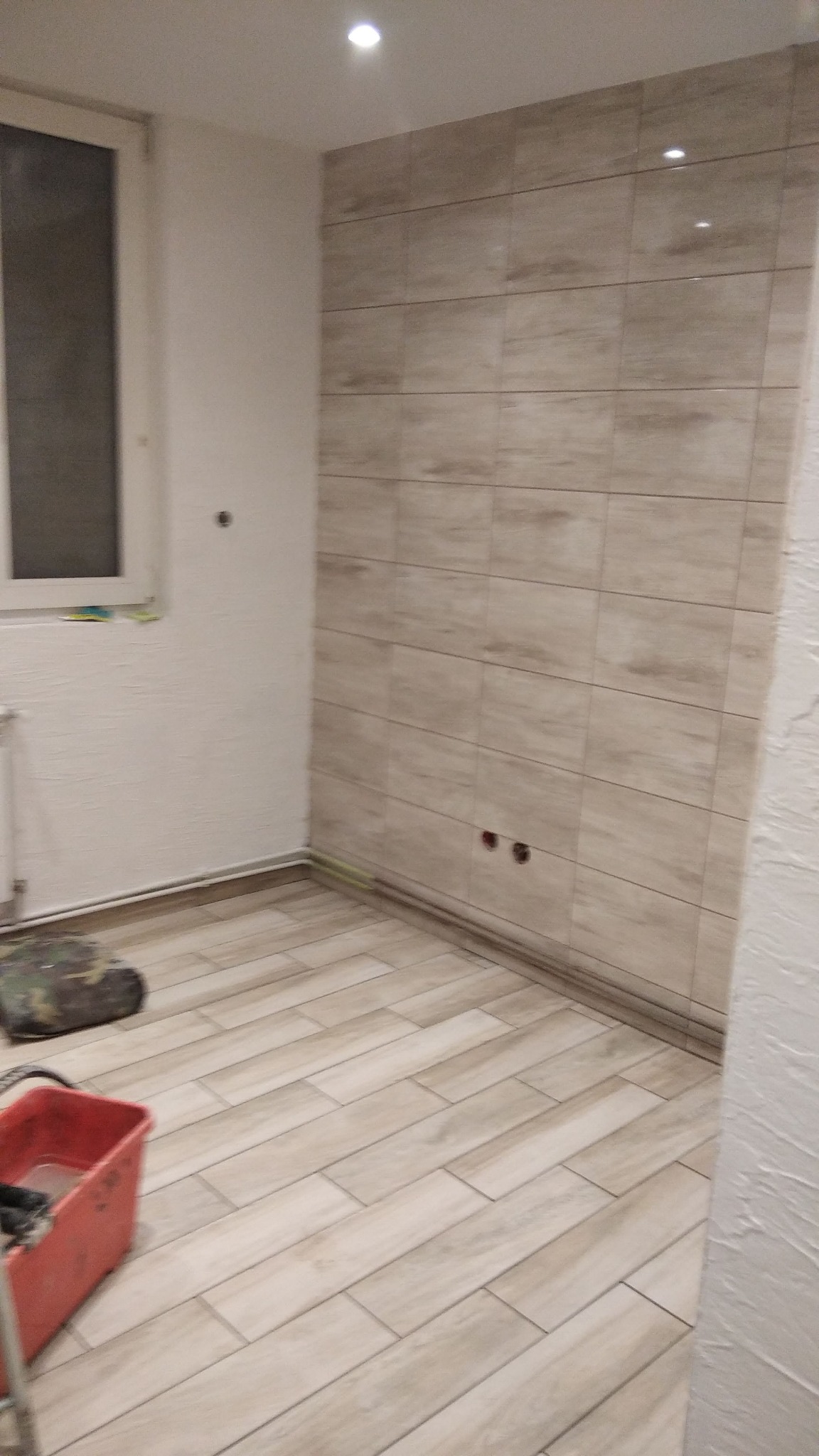 Wykończona łazienka z beżowymi płytkami imitującymi drewno na ścianie i podłodze, widoczny czerwony pojemnik i narzędzia malarskie, przygotowane do dalszych prac wykończeniowych.