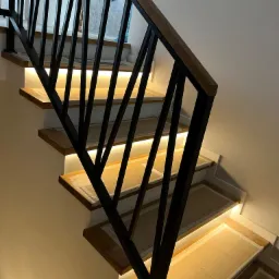 Schody drewniane z czarną balustradą i podświetleniem LED pod stopniami, zabezpieczone kartonem przed uszkodzeniami podczas prac wykończeniowych.