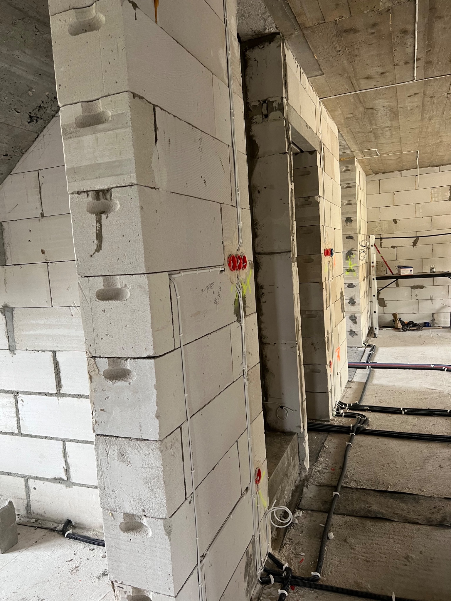 Wnętrze w trakcie budowy z widocznymi ścianami z białych bloków, instalacją elektryczną w peszlach na podłodze i puszkami elektrycznymi w ścianach.
