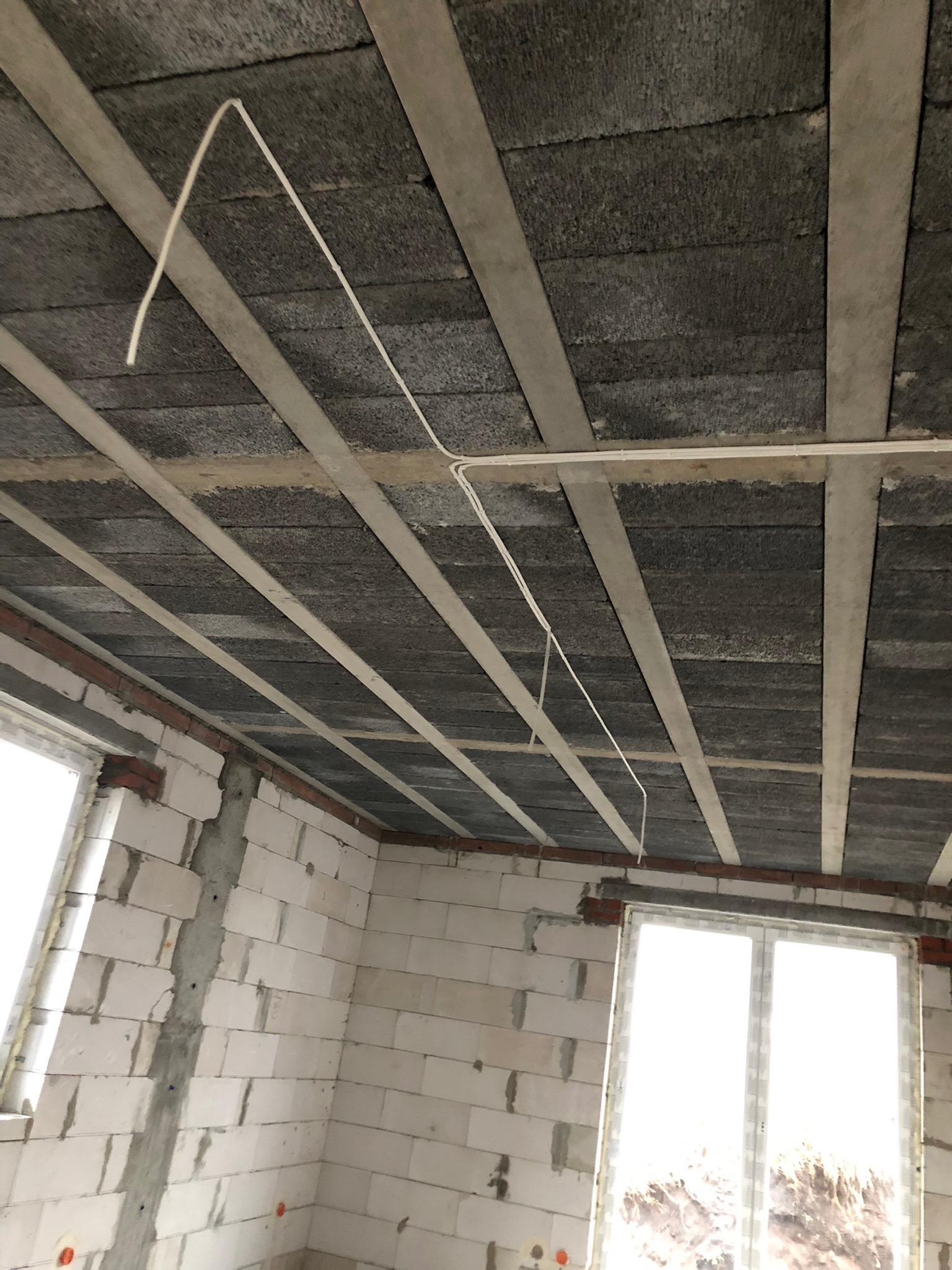 Surowy stan budynku w trakcie prac: widoczne przewody elektryczne pod sufitem z betonowych płyt, ściany z białych bloczków, okna bez parapetów.