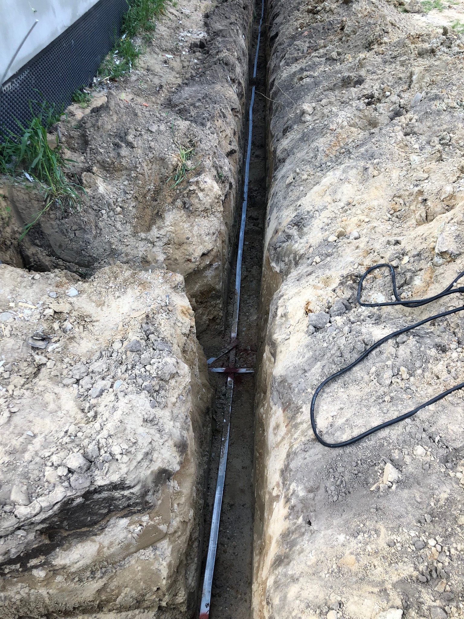 Wykop w ziemi z ułożoną metalową rurą i czarnym kablem elektrycznym, widoczne ślady świeżo wykonanych prac ziemnych.