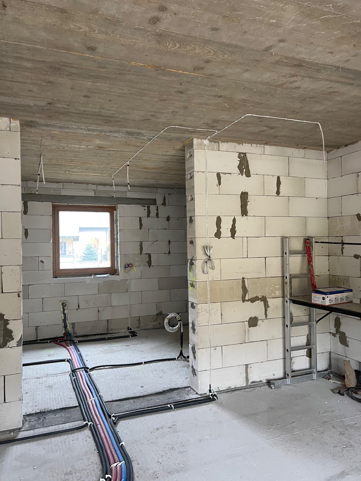 Wnętrze w trakcie prac instalacyjnych: widoczne przewody elektryczne prowadzone po suficie i podłodze, ściany z betonu komórkowego z widocznymi zaprawami, drabina oparta o ścianę, surowy betonowy...