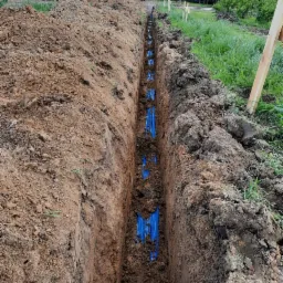 Wykopany rów z ułożonymi, niebieskimi rurami instalacyjnymi, widok z góry wzdłuż wykopu, ziemia po bokach, częściowo zasypana, w tle budynek gospodarczy i samochód dostawczy.