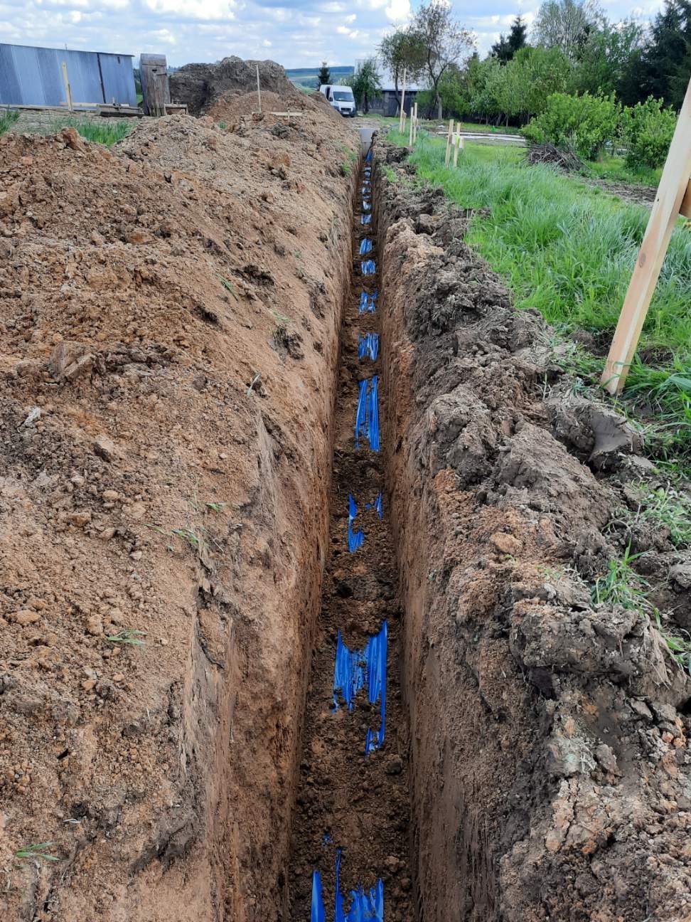 Wykopany rów z ułożonymi, niebieskimi rurami instalacyjnymi, widok z góry wzdłuż wykopu, ziemia po bokach, częściowo zasypana, w tle budynek gospodarczy i samochód dostawczy.